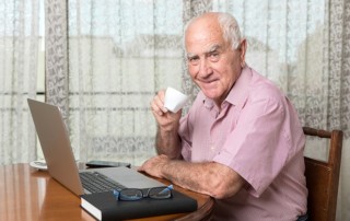 Older man smiling while drinking coffee or tea and working on his computer
