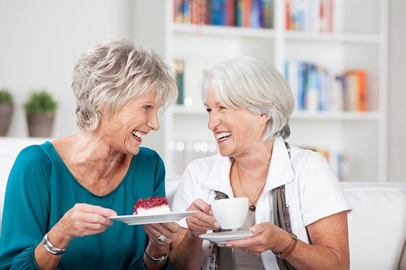 Old friends laughing together while drinking coffee and eating a pastry in a living room.