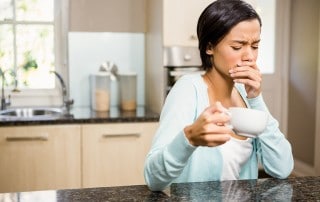 A woman holding her mouth with one hand and a cup of coffee in the other, while in her kitchen.